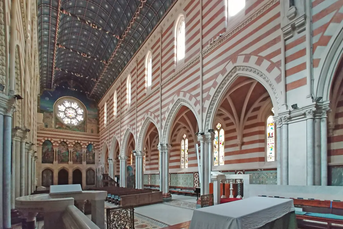 The side of a church interior seen from a gallery, with striped walls, a star-patterned blue ceiling, pointed arches and a tiled floor in red and green.