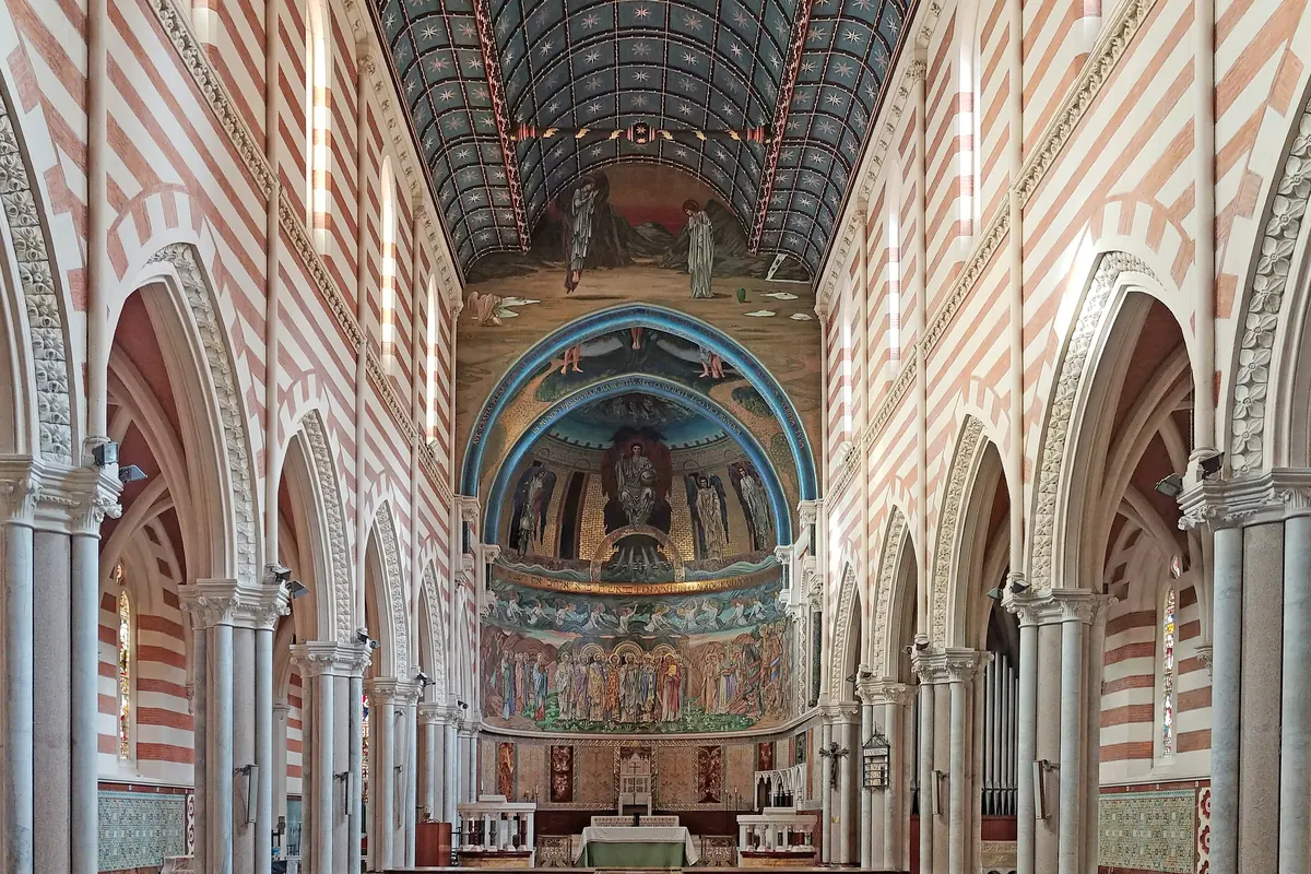 A long nave with walls of red and white stripes and pointed arches, rows of wooden pews on a patterned floor, and a mosaic-covered apse at the far end.