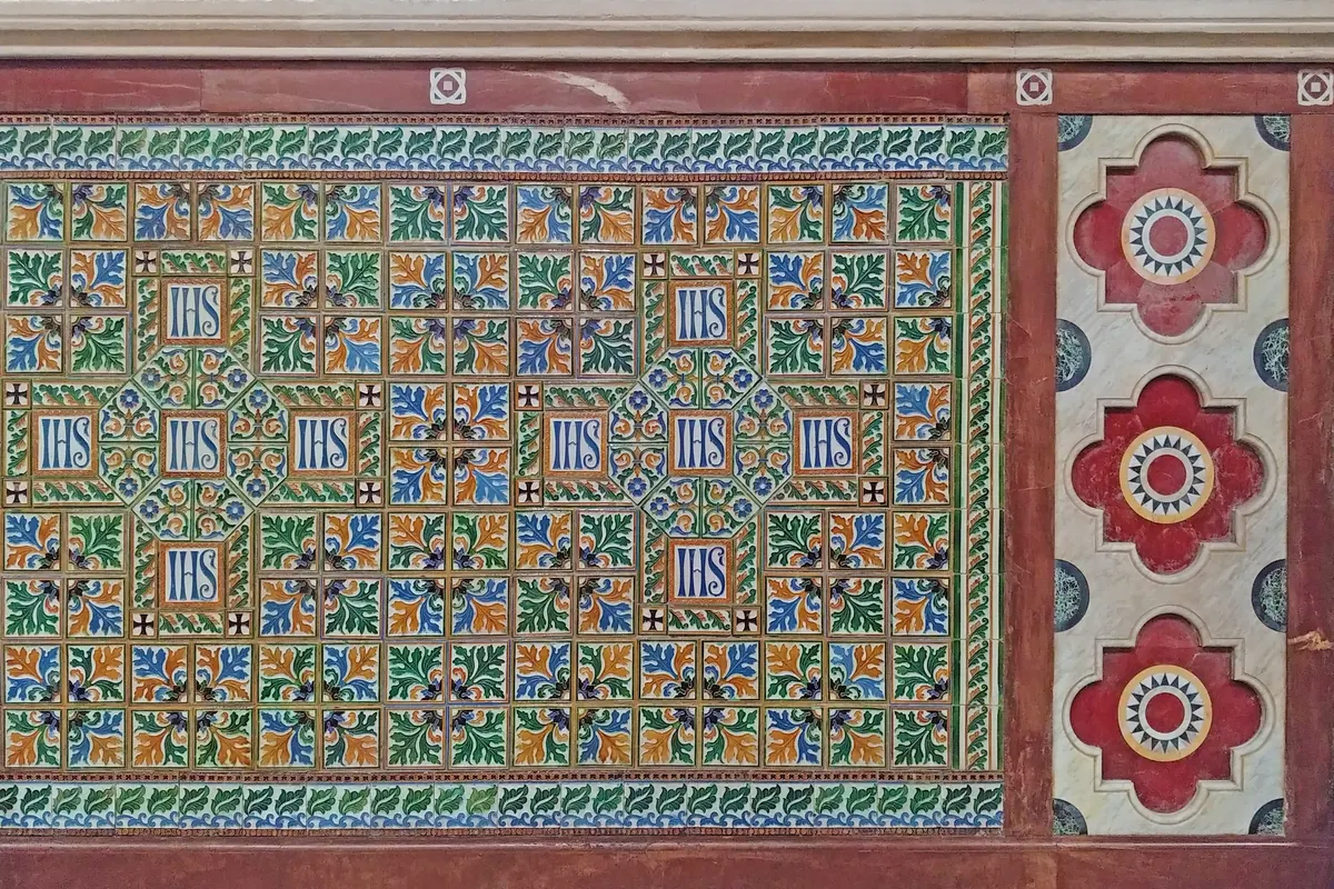 A wall panel of green, blue and orange glazed tiles forming a repeating pattern with the letters IHS, next to a marble panel with three red quatrefoil medallions.